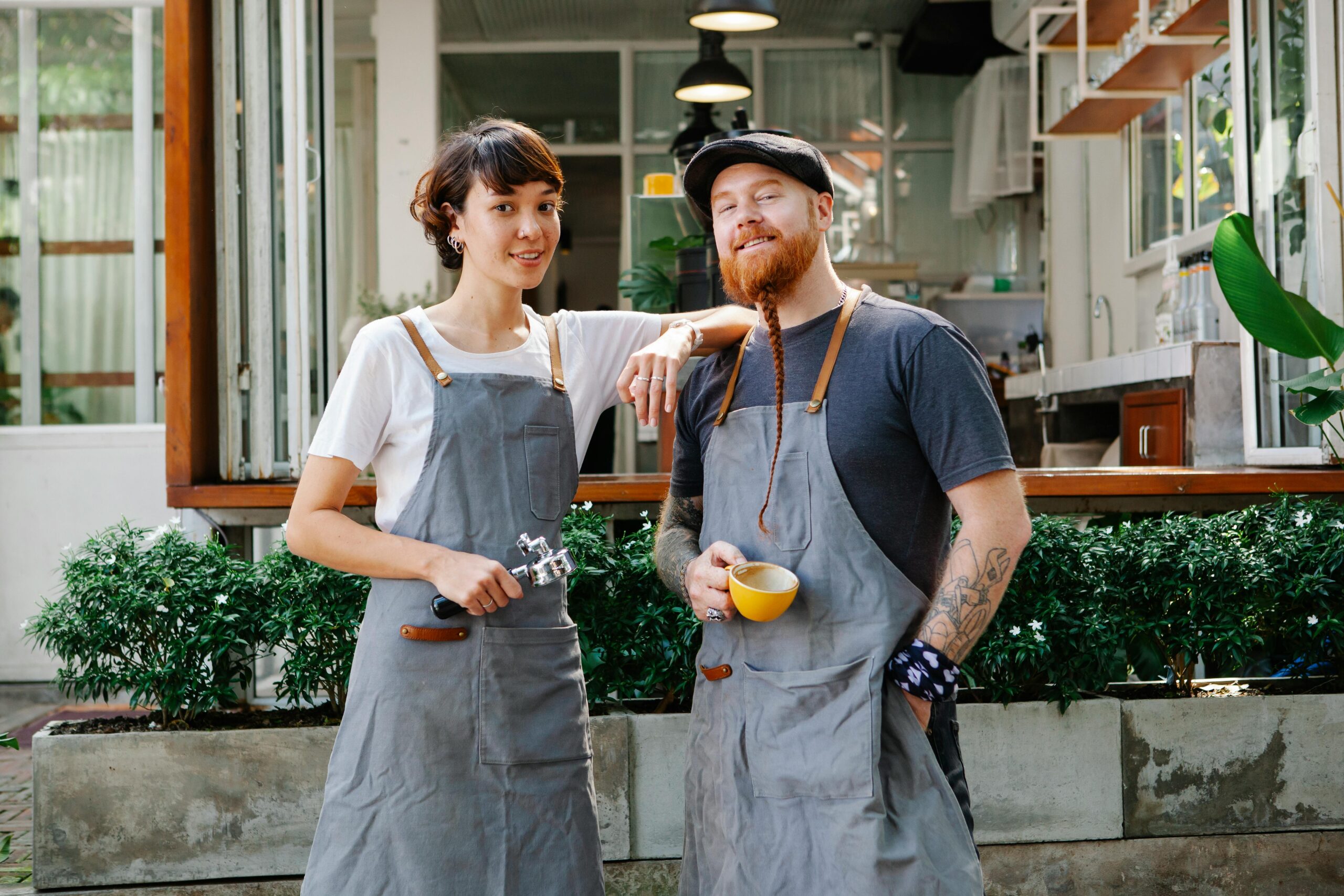 Two baristas in aprons smiling at their cozy outdoor coffee shop.